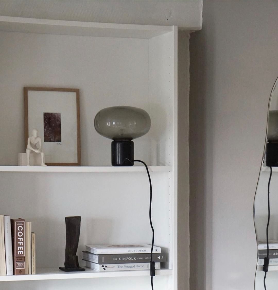 An unlit Clérachic table lamp on a white bookshelf, set between a framed portrait, a small figurine and stacked books.