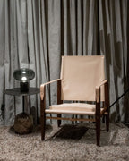 The Clérachic table lamp on a round black side table beside a tan leather armchair, against a heavy curtain backdrop.