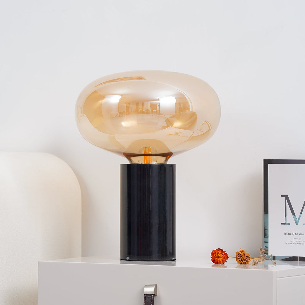 The amber-glass Clérachic table lamp on a white cabinet, with dried flowers and a framed print beside its black base.