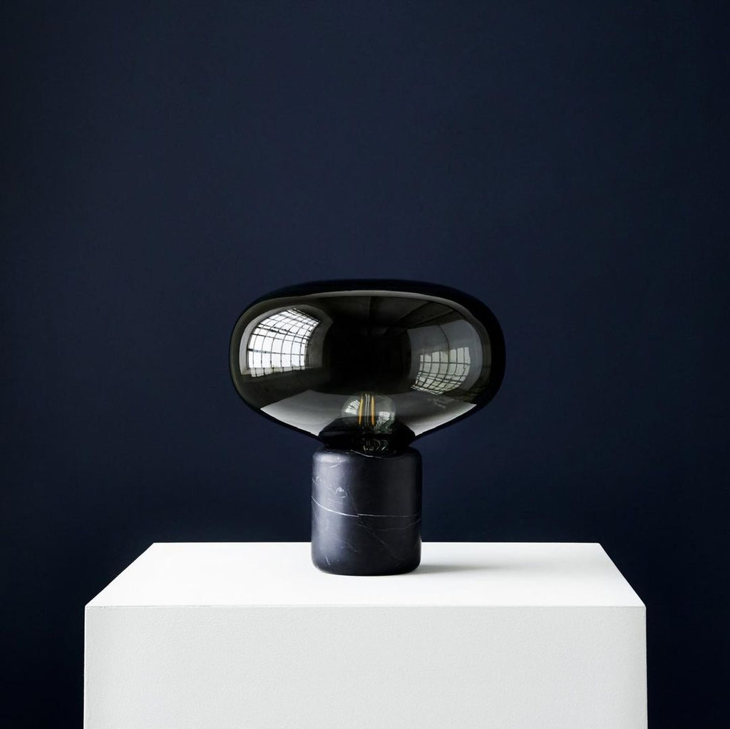 The Clérachic table lamp on a white plinth against a dark navy wall, its glass dome catching a single highlight.