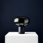 The Clérachic table lamp on a white plinth against a dark navy wall, its glass dome catching a single highlight.