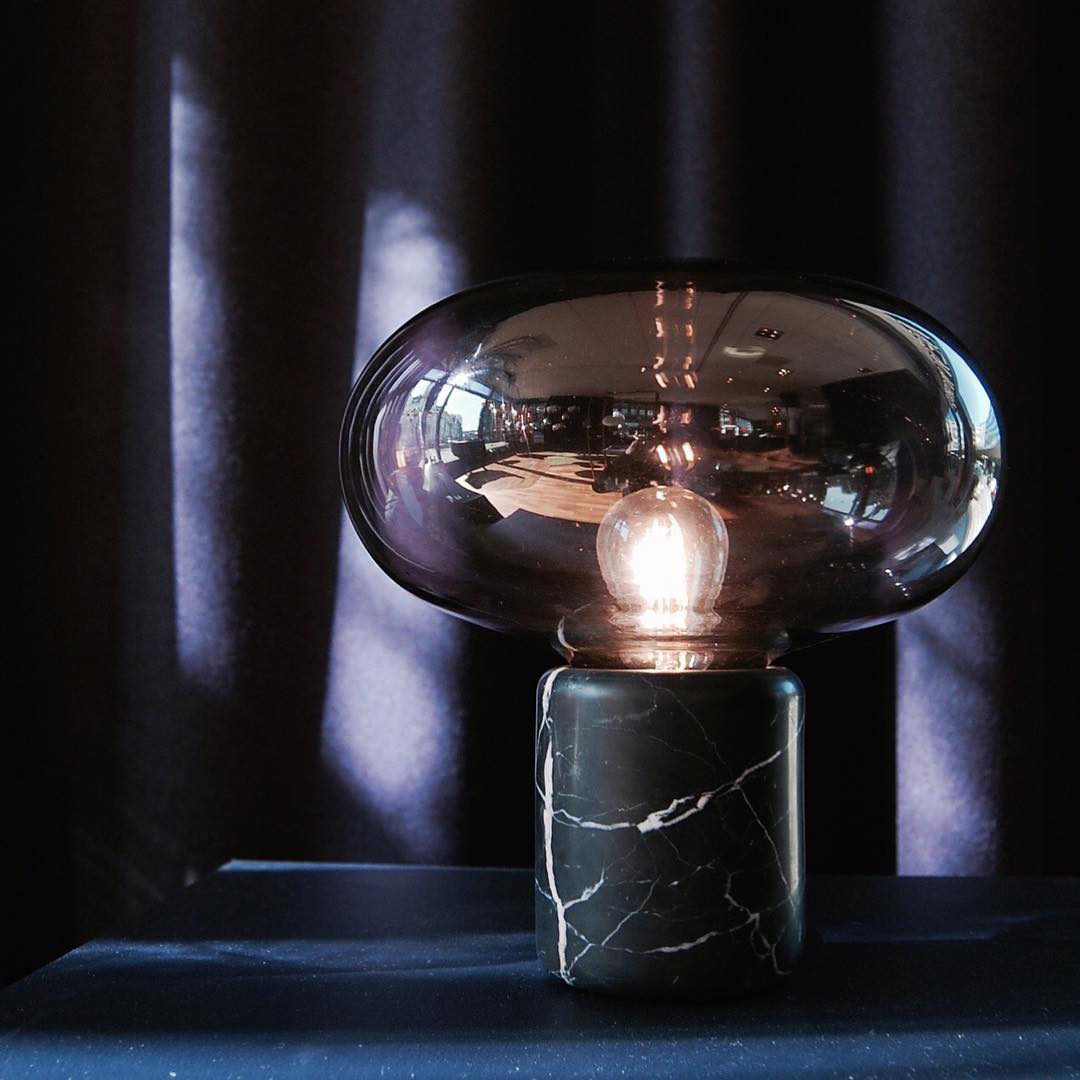 The lit Clérachic table lamp against a dark curtain backdrop, its glass dome glowing warmly above the marble base.