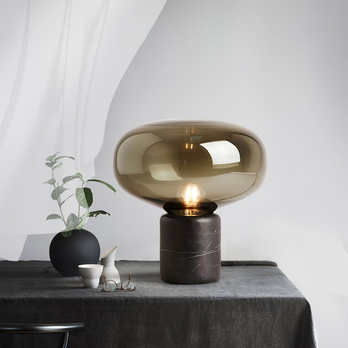 The lit amber-glass Clérachic table lamp on a grey tablecloth, beside a small black vase and ceramic pieces.