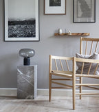 An unlit Clérachic table lamp on a grey stone pedestal, with wooden chairs and framed artwork in the room behind it.