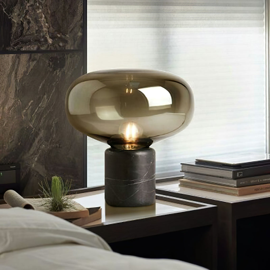The lit amber-glass Clérachic table lamp on a table by a curtained window, with potted plants nearby.