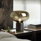 The lit amber-glass Clérachic table lamp on a table by a curtained window, with potted plants nearby.