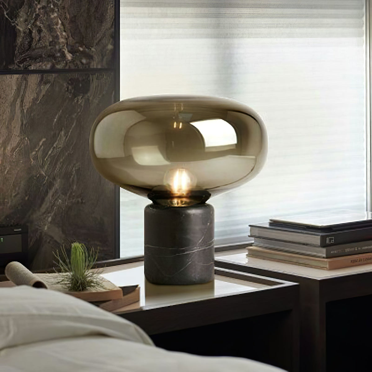 The lit amber-glass Clérachic table lamp on a table by a curtained window, with potted plants nearby.
