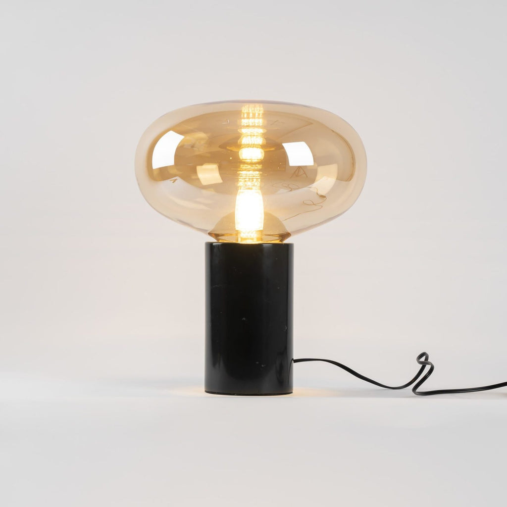 A studio shot of the amber-glass Clérachic table lamp on white, its cord looped to one side of the marble base.