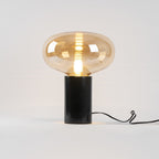 A studio shot of the amber-glass Clérachic table lamp on white, its cord looped to one side of the marble base.