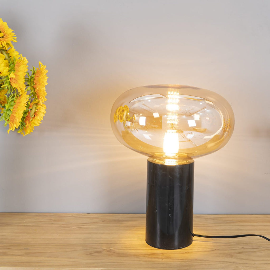 The lit amber-glass Clérachic table lamp on a wood table beside cut sunflowers, its marble base grounding the scene.