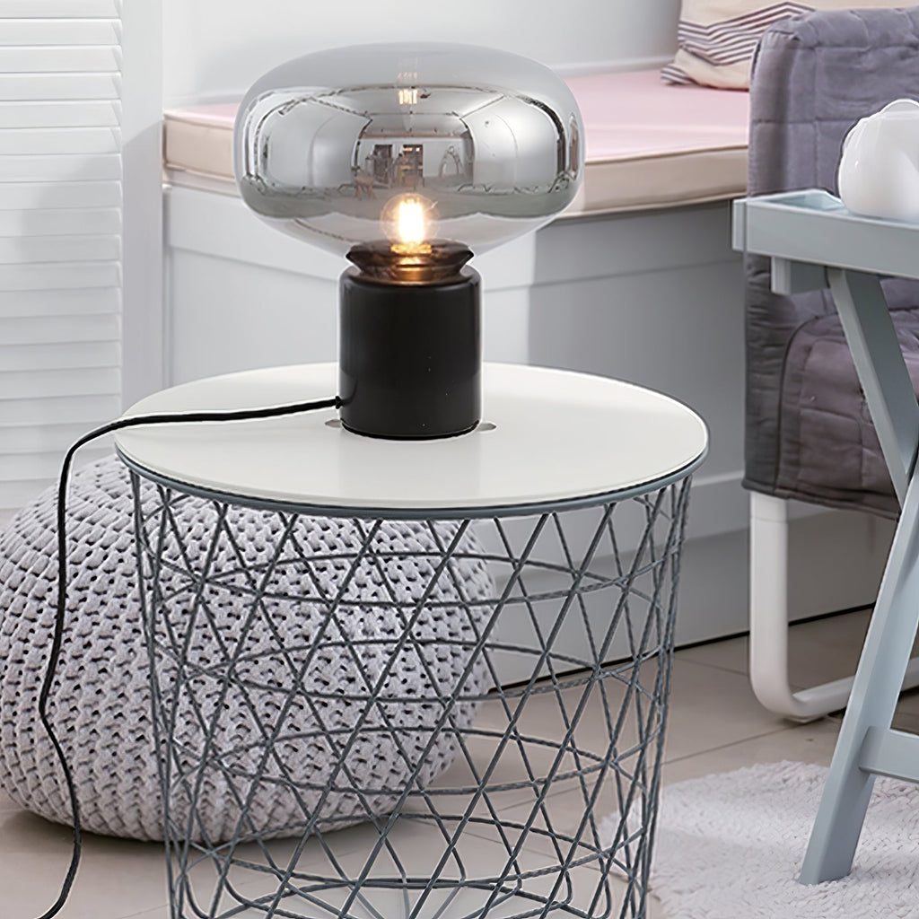 The lit Clérachic table lamp on a white wire basket side table, a soft blanket blurred in the background.