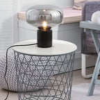 The lit Clérachic table lamp on a white wire basket side table, a soft blanket blurred in the background.