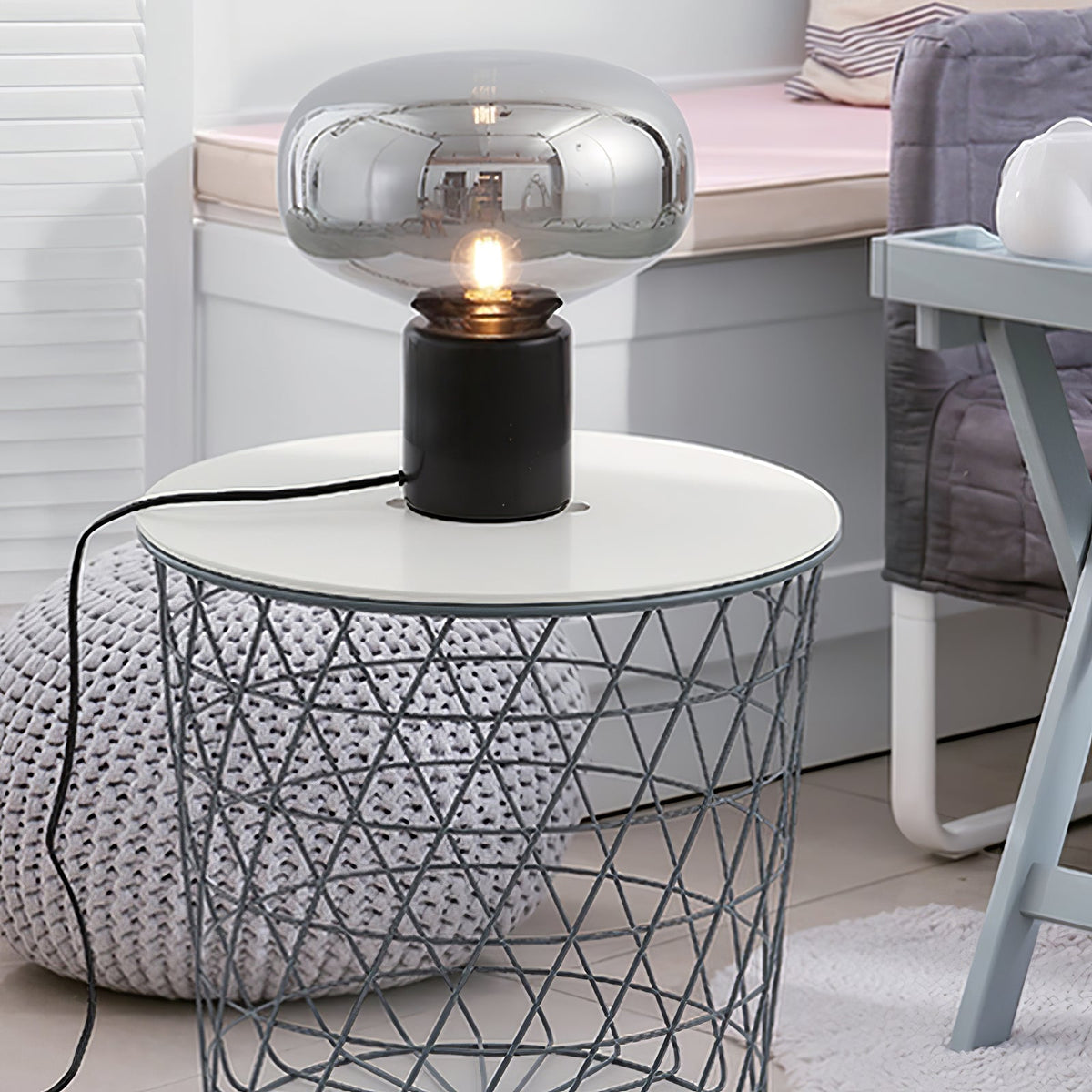 The lit Clérachic table lamp on a white wire basket side table, a soft blanket blurred in the background.