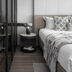 The Clérachic table lamp on a round nightstand beside a bed dressed in white linen and a grey faux-fur throw.