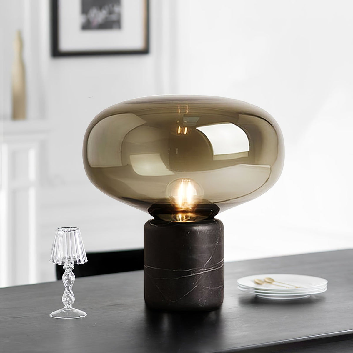 The Clérachic table lamp on a dark table beside a miniature glass lamp ornament and a small stack of plates.