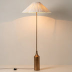 Switched on and glowing against a plain wall, a Clérachic pleated white cone-shade floor lamp rests on its brass bottle base.