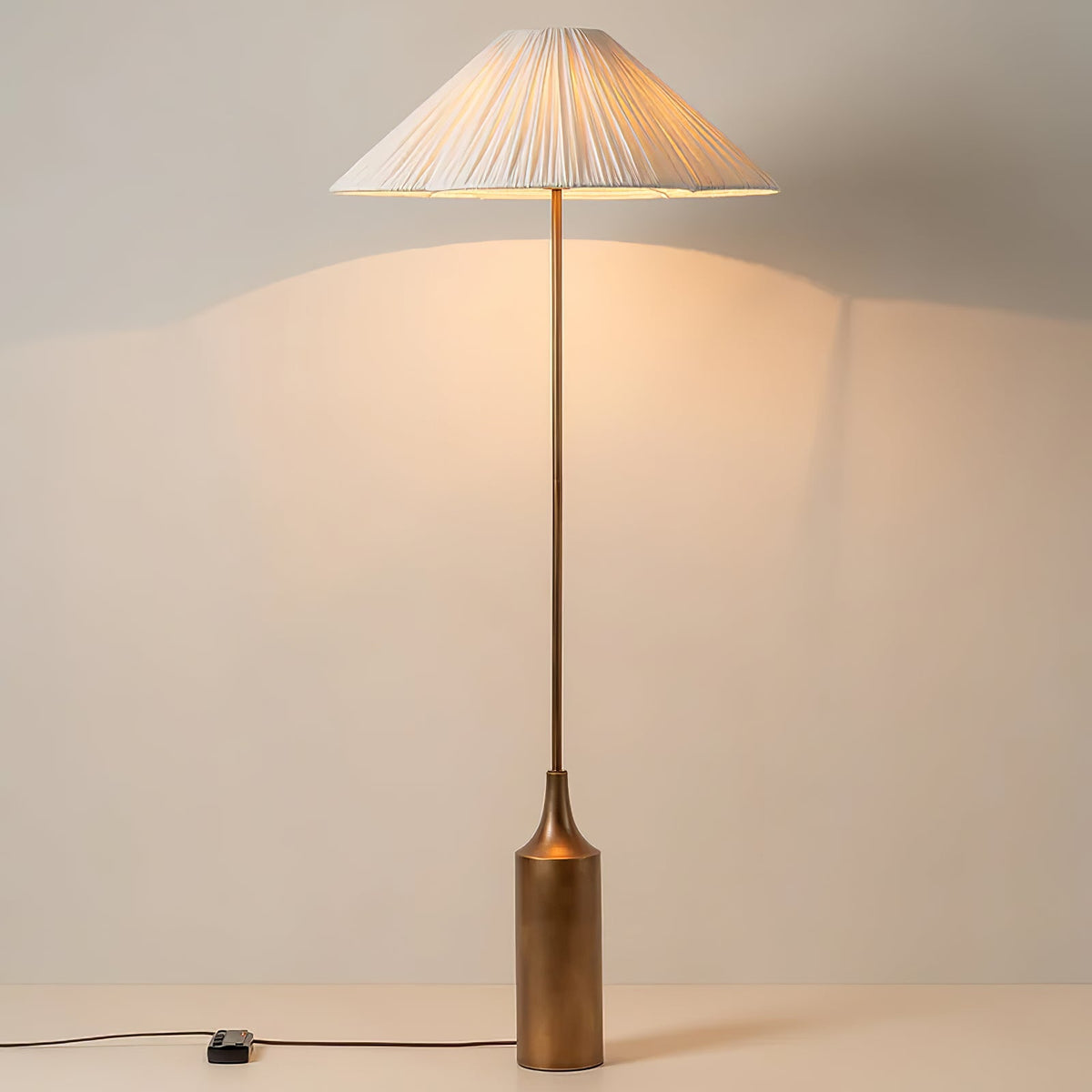 Switched on and glowing against a plain wall, a Clérachic pleated white cone-shade floor lamp rests on its brass bottle base.