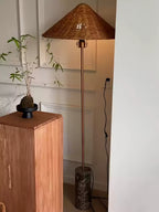 In a corner beside a wooden plinth and potted plant, a Clérachic rattan cone-shade floor lamp stands on its marble base.