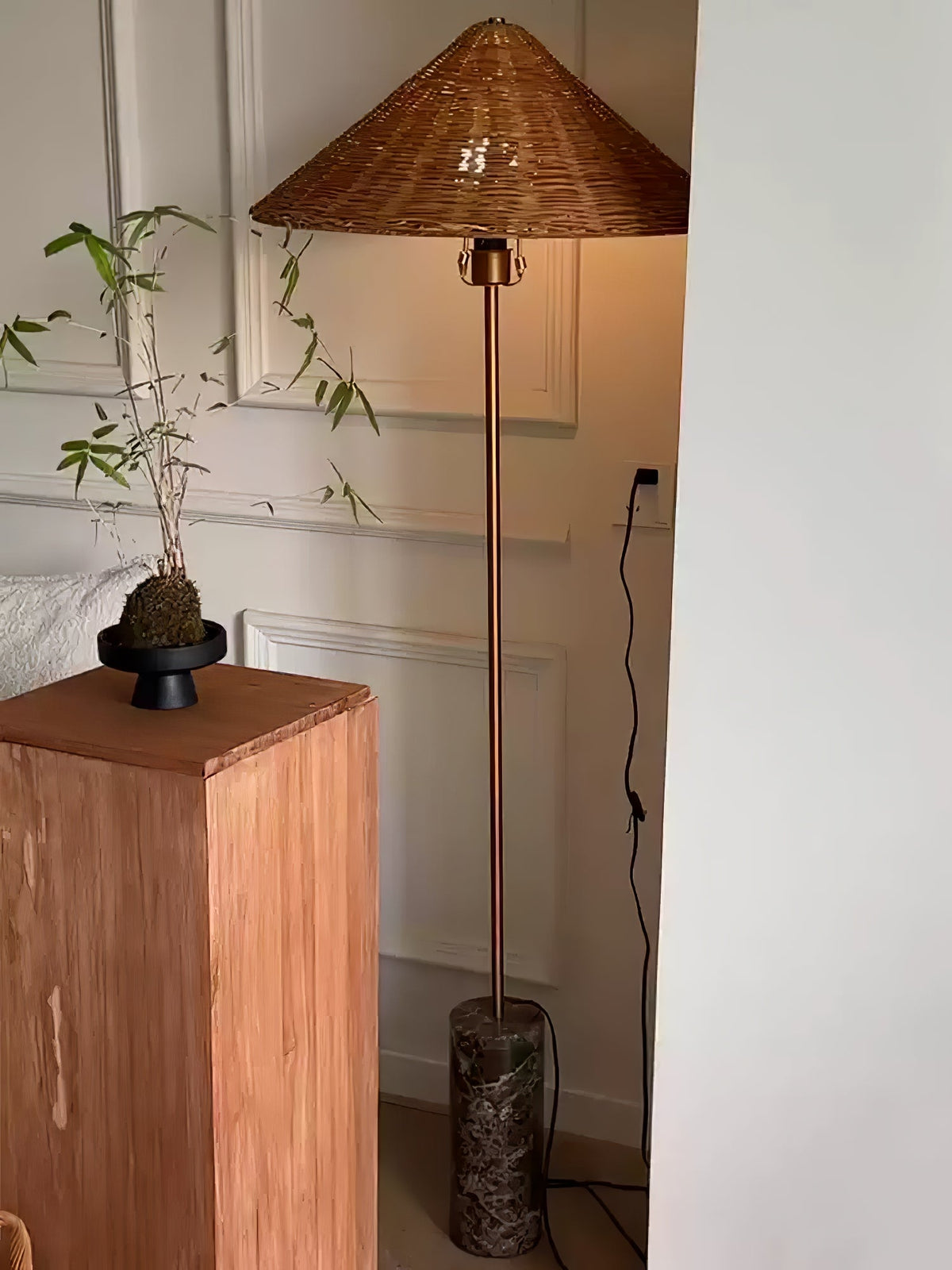 In a corner beside a wooden plinth and potted plant, a Clérachic rattan cone-shade floor lamp stands on its marble base.