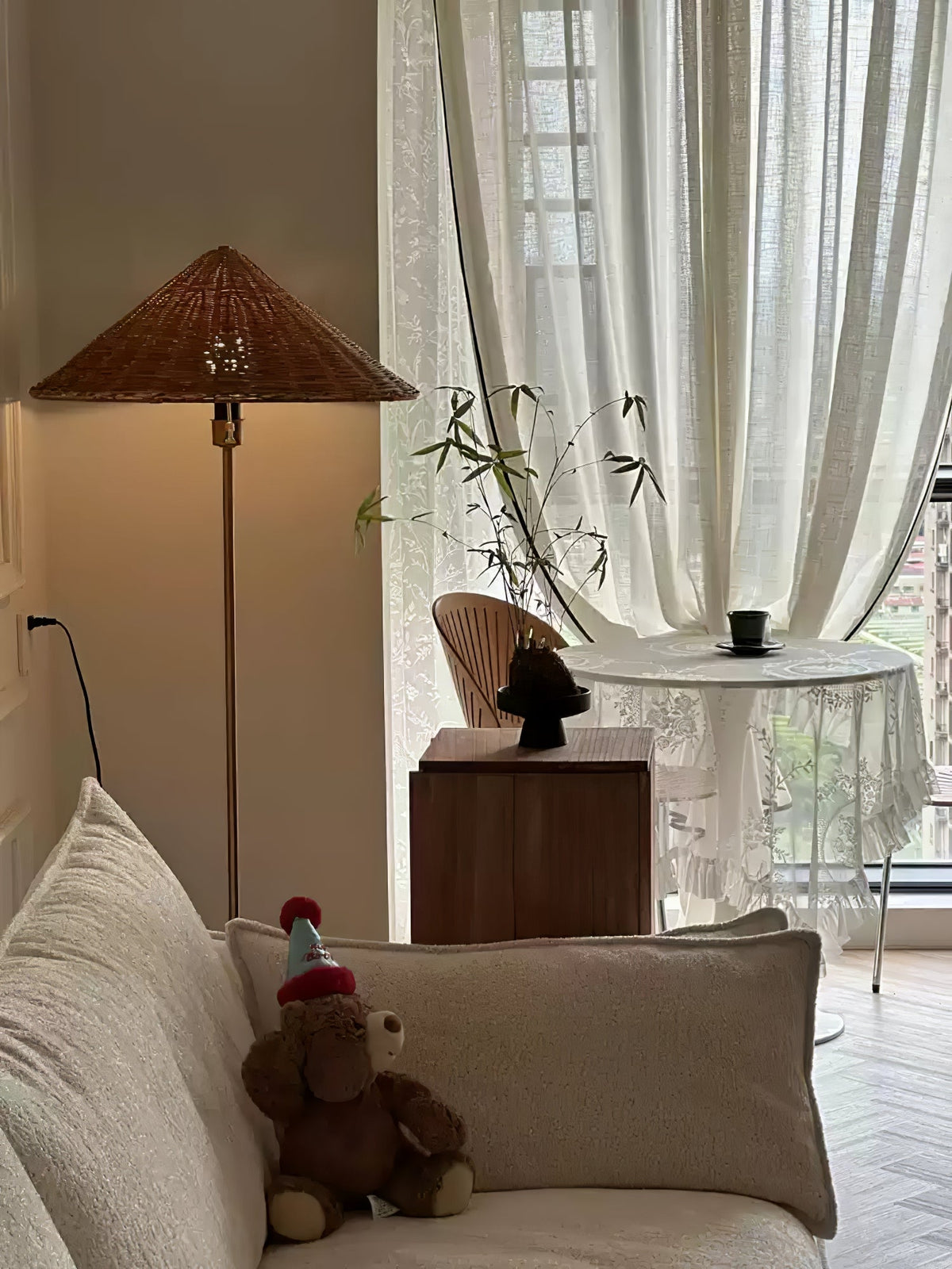 Warm light spreads near sheer curtains as this Clérachic rattan cone-shade floor lamp glows beside a living room window.