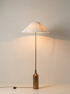 Clérachic pleated white cone-shade floor lamp on a tall brass pole, switched on and casting warm light up a plain wall in the corner.