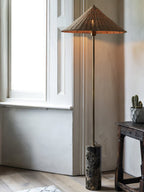 Clérachic rattan cone-shade floor lamp glowing beside a window, its warm light spreading across the wall in a sunlit corner.