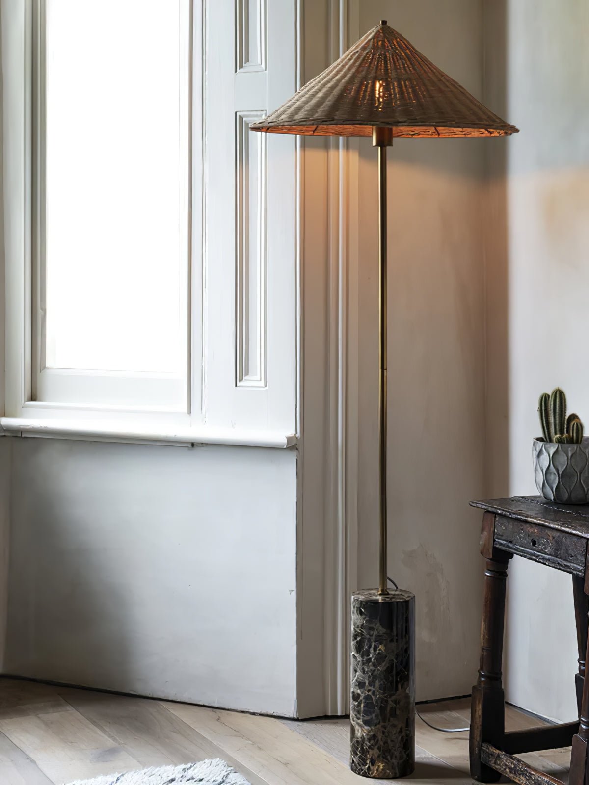 Clérachic rattan cone-shade floor lamp glowing beside a window, its warm light spreading across the wall in a sunlit corner.
