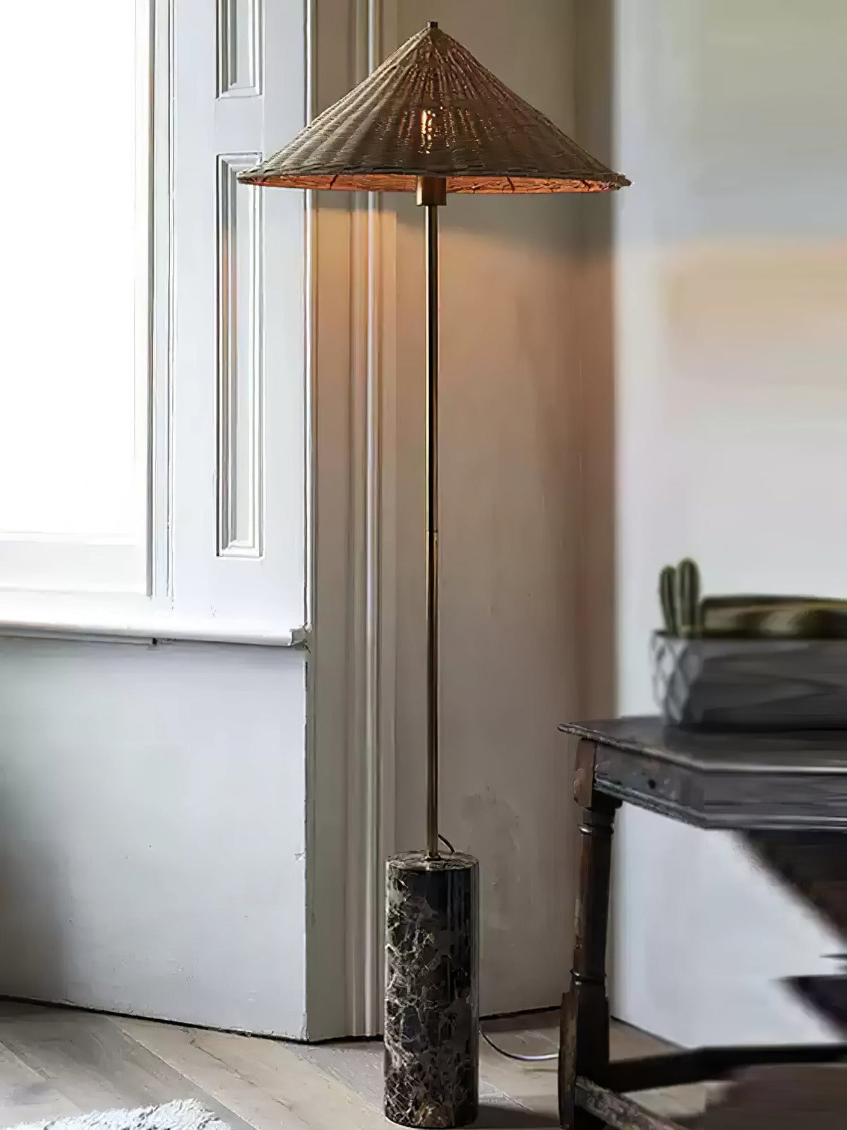 Clérachic rattan cone-shade floor lamp beside a small round table, with its dark marble base and brass stem in daylight.