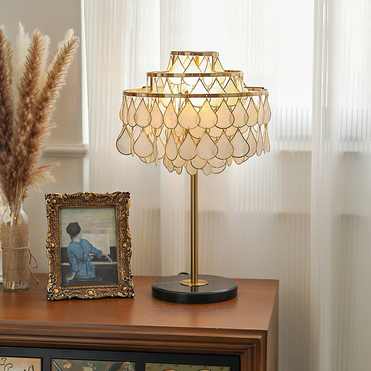 Clérachic table lamp lit on a dark wood sideboard beside dried pampas grass and a gilt-framed vintage photograph.