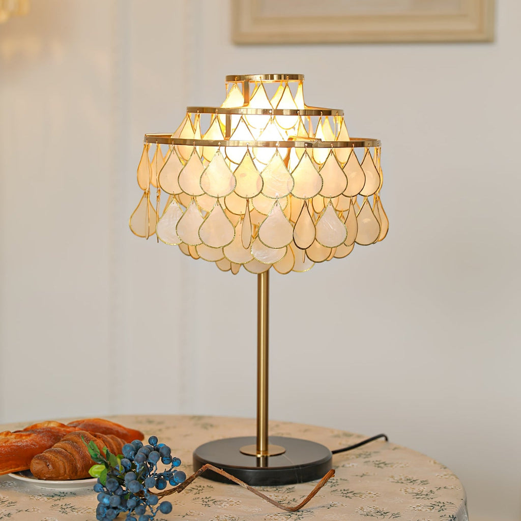 Clérachic teardrop shell table lamp glowing on a round table set with a china plate of croissants and blueberries.