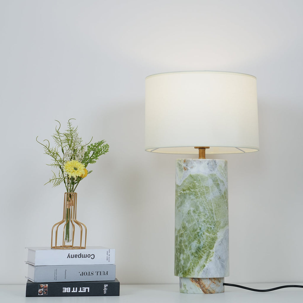 Clérachic table lamp beside a flower-topped book stack, its white drum shade lit to a soft warm tone.