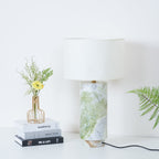 Clérachic table lamp in a wider crop, showing the full drum shade above the green marble base and fern fronds nearby.
