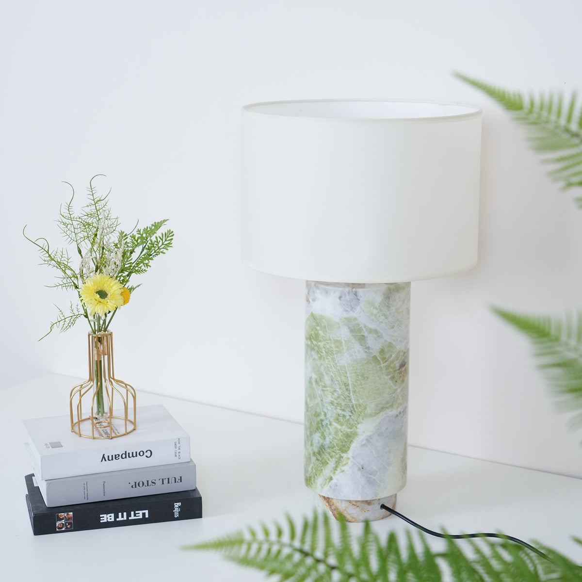 Clérachic table lamp framed closely by fern fronds on both sides, with the flower vase and books to its left.