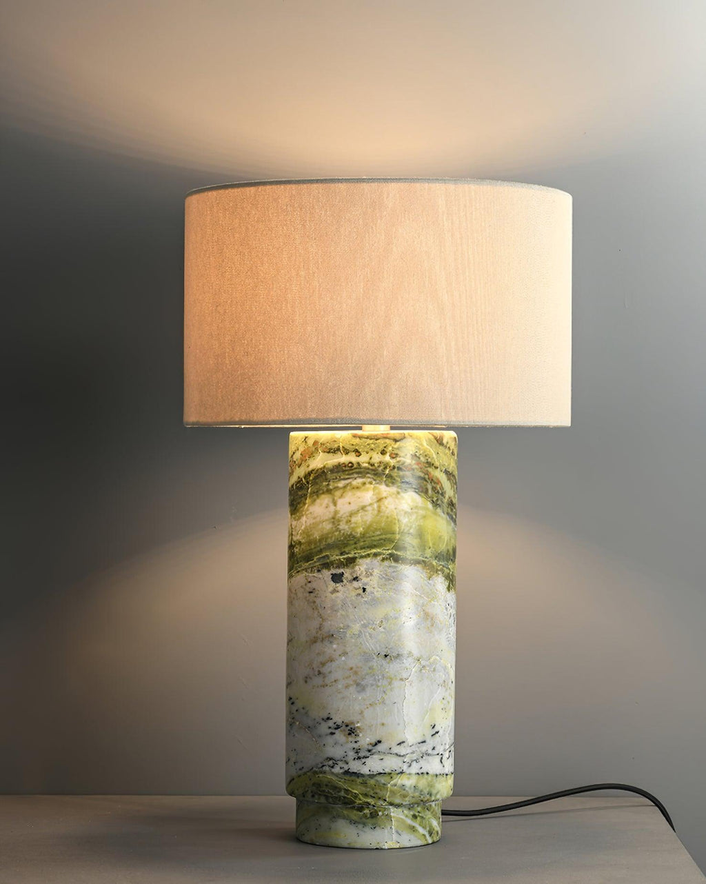 Clérachic table lamp on a grey background, its lit white drum shade casting a warm glow across the wall behind it.