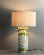Clérachic table lamp on a grey background, its lit white drum shade casting a warm glow across the wall behind it.
