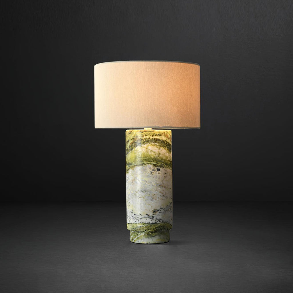 Clérachic table lamp against a black background, its white drum shade lit warmly to highlight the green marble base.