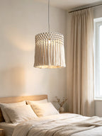 Above a low bed with cream linens, this Clérachic pendant lights a bright bedroom corner by a curtained window.