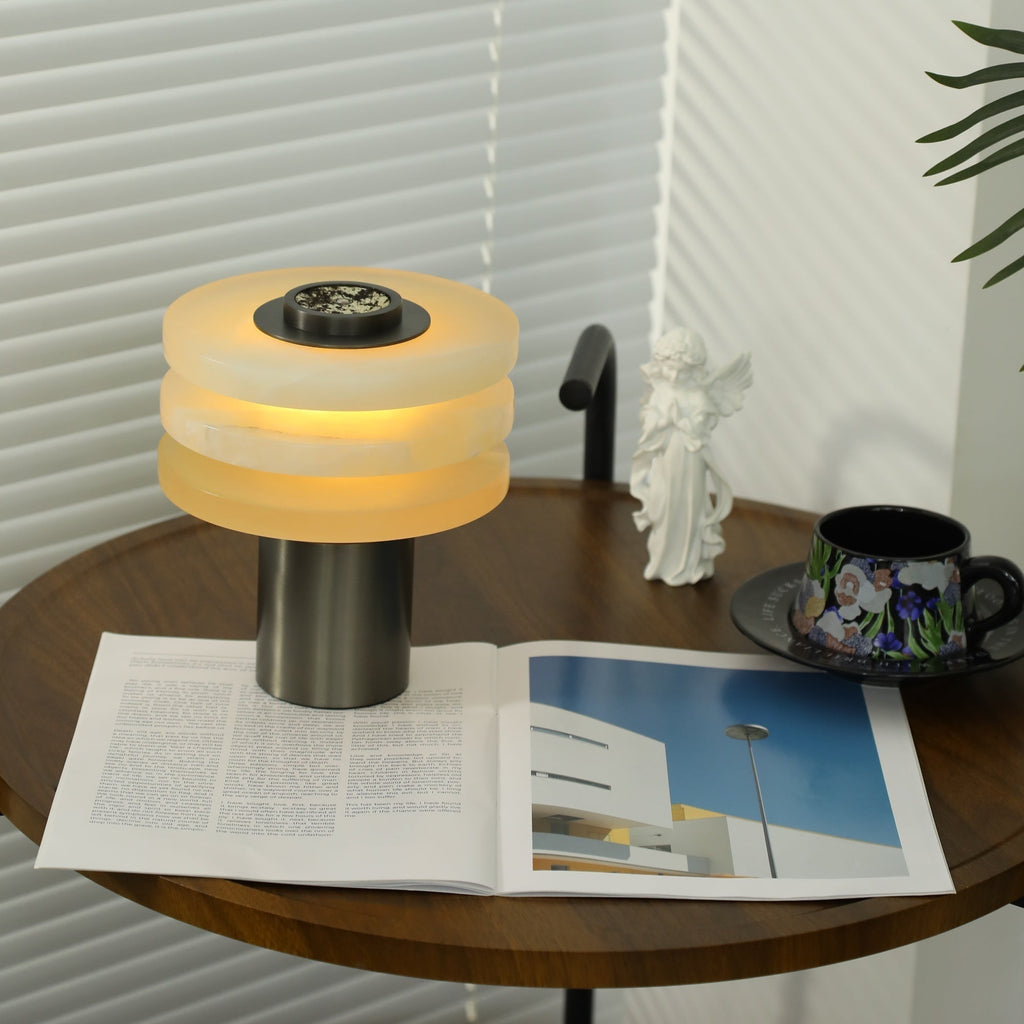 Wide lifestyle shot of the lit alabaster lamp on a walnut table beside a floral teacup and open magazine