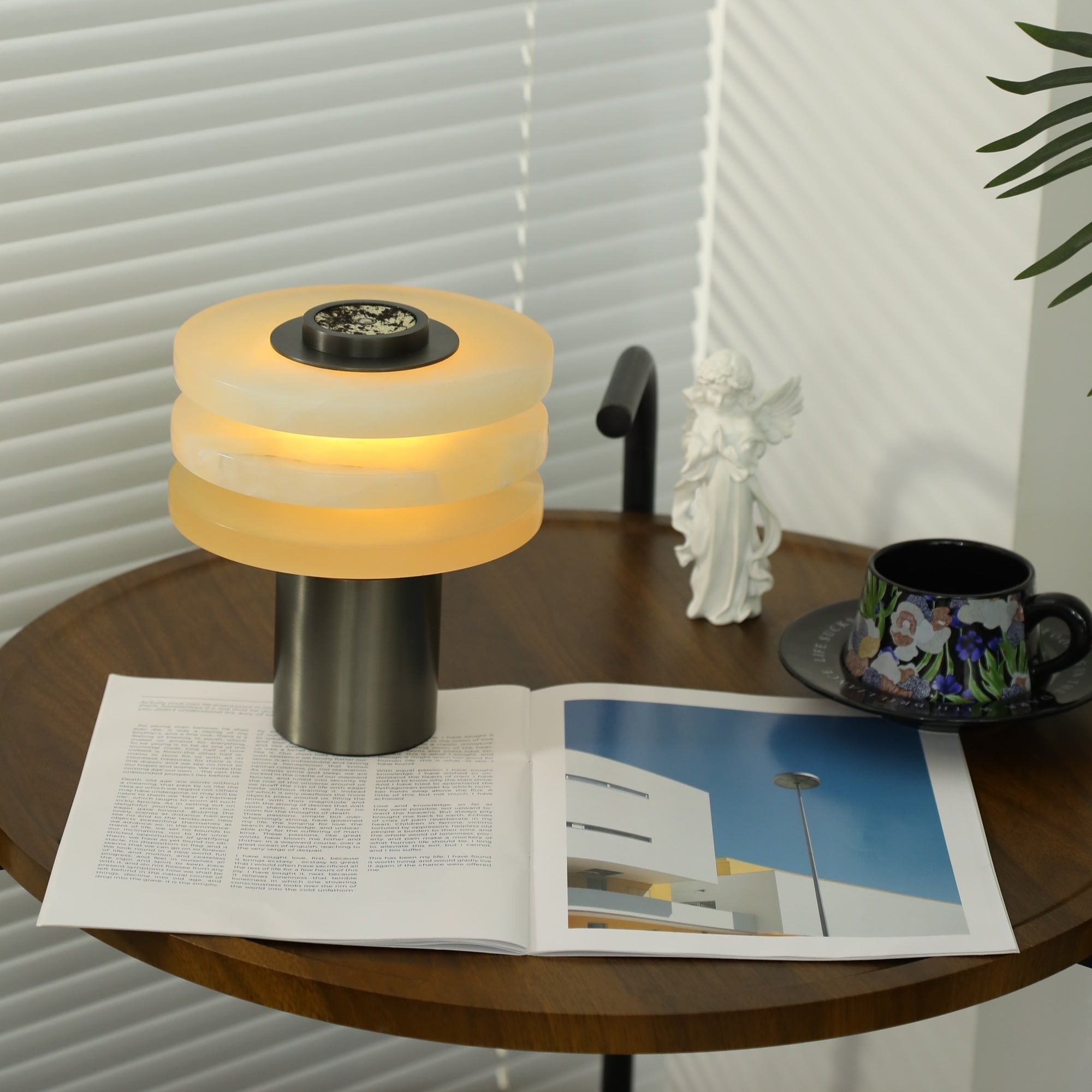 Wide lifestyle shot of the lit alabaster lamp on a walnut table beside a floral teacup and open magazine