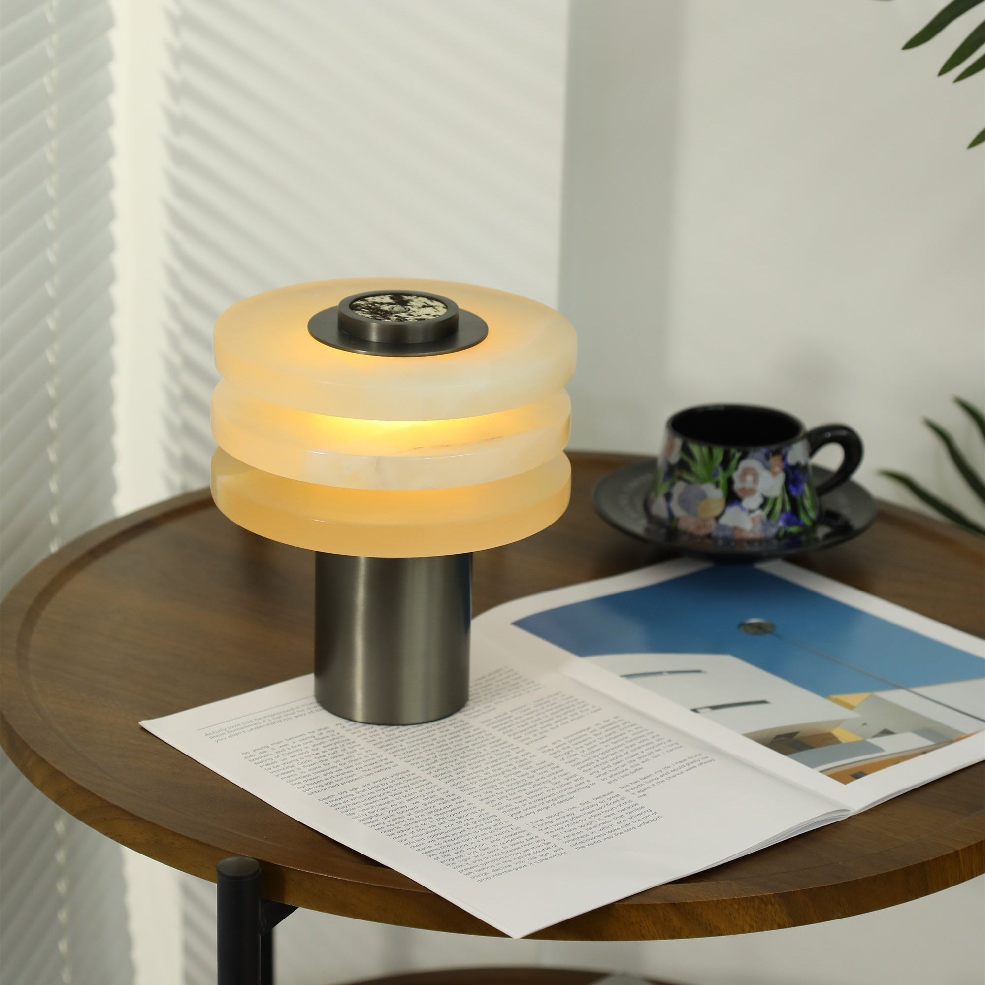 The lit lamp resting on an open magazine, its stacked alabaster shade pooling warm light on the table