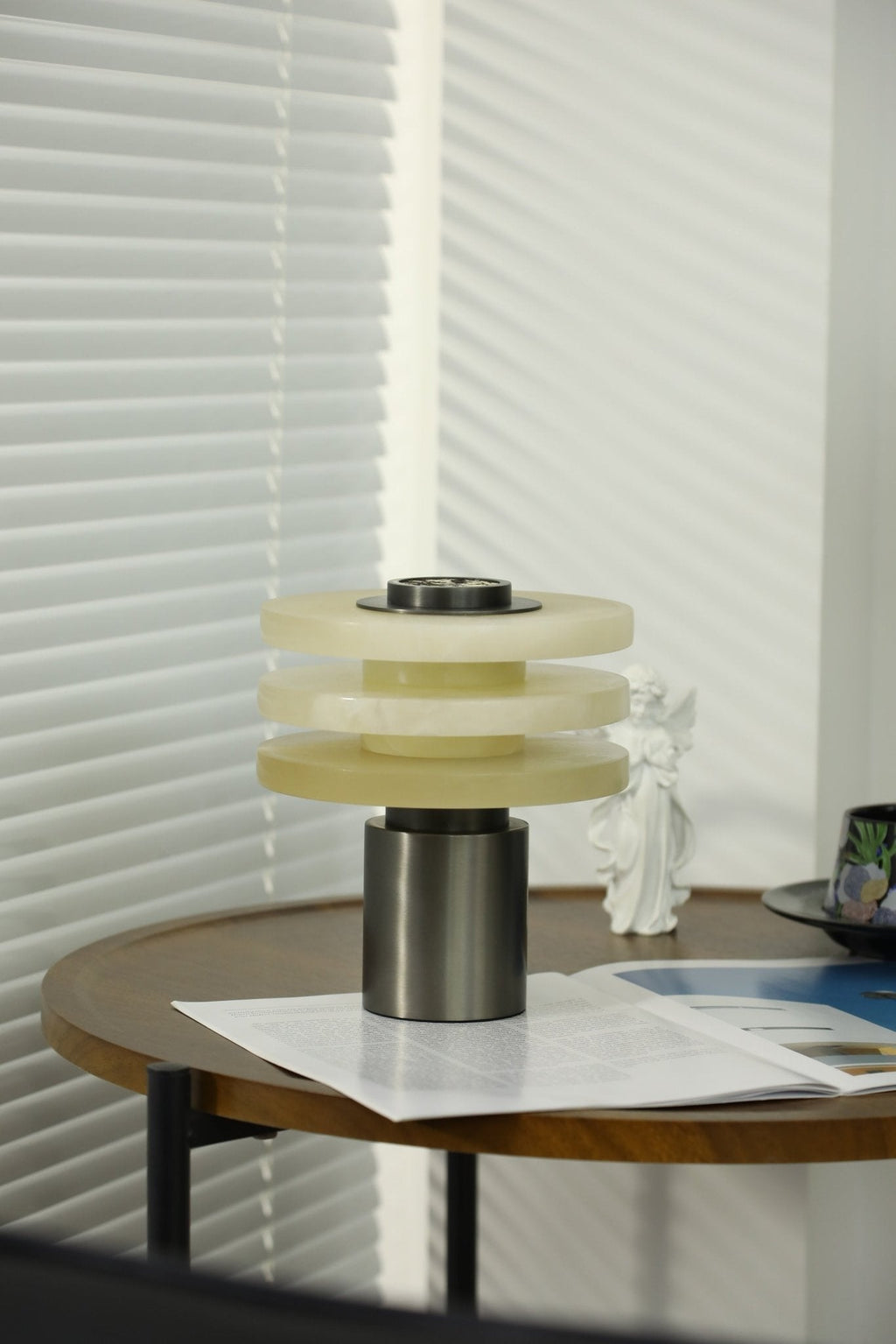 Tall view of the unlit alabaster lamp on a walnut side table, daylight striping the white blinds behind