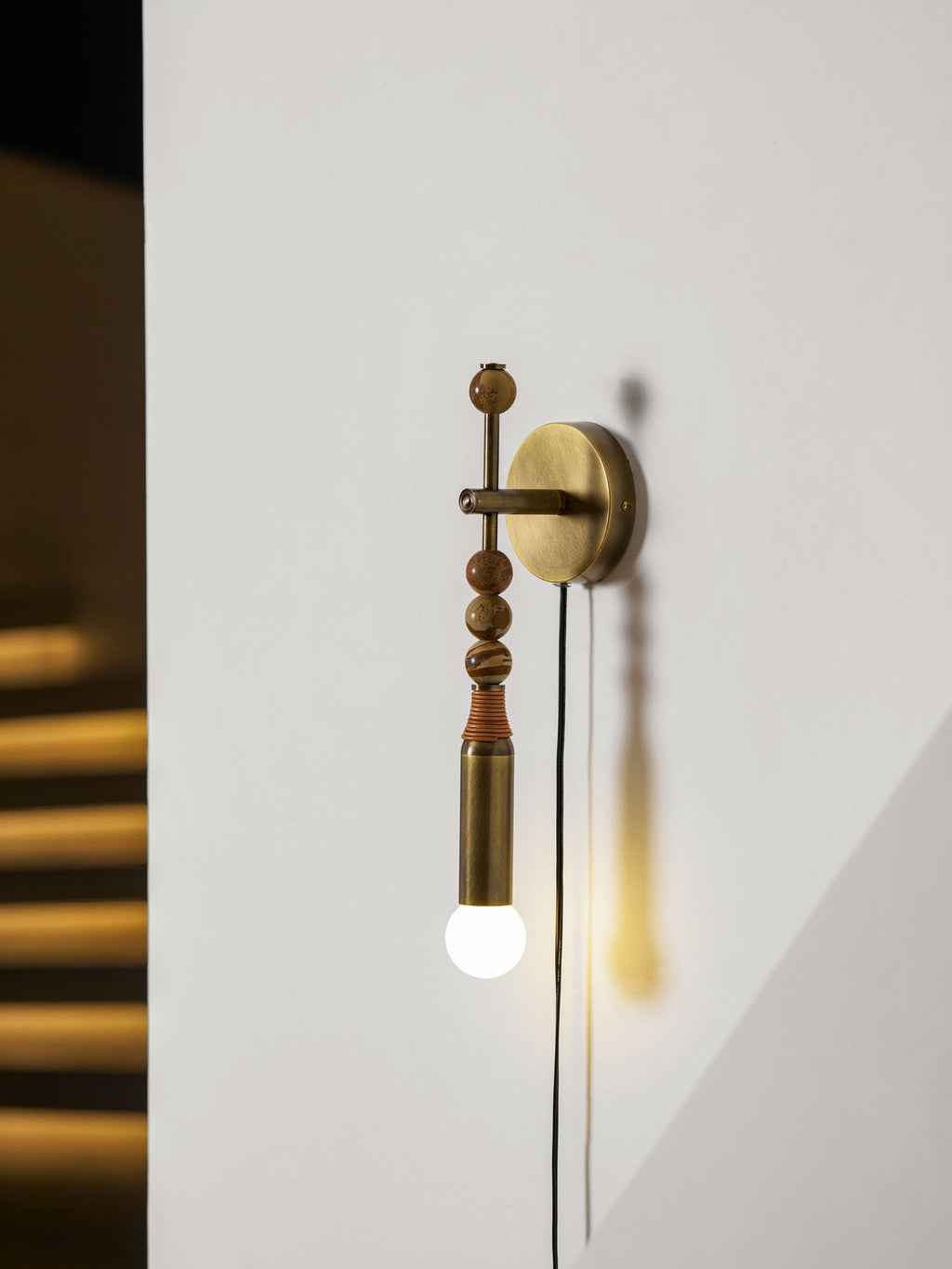 Clérachic plug-in wall lamp on a plain wall beside a dark staircase, wood beads above a lit glass globe.