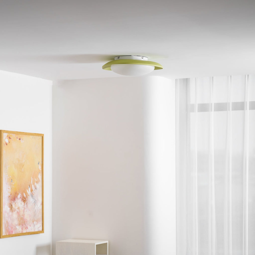 Close beneath the ceiling near sheer curtains, the green rim of this Clérachic ceiling light is visible against the wall.