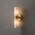 In brushed brass, this Clérachic wall light hangs on a plain grey wall, its two alabaster tubes glowing softly.