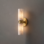 In brushed brass, this Clérachic wall light hangs on a plain grey wall, its two alabaster tubes glowing softly.