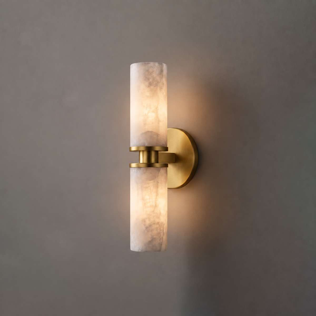 In brushed brass, this Clérachic wall light hangs on a plain grey wall, its two alabaster tubes glowing softly.