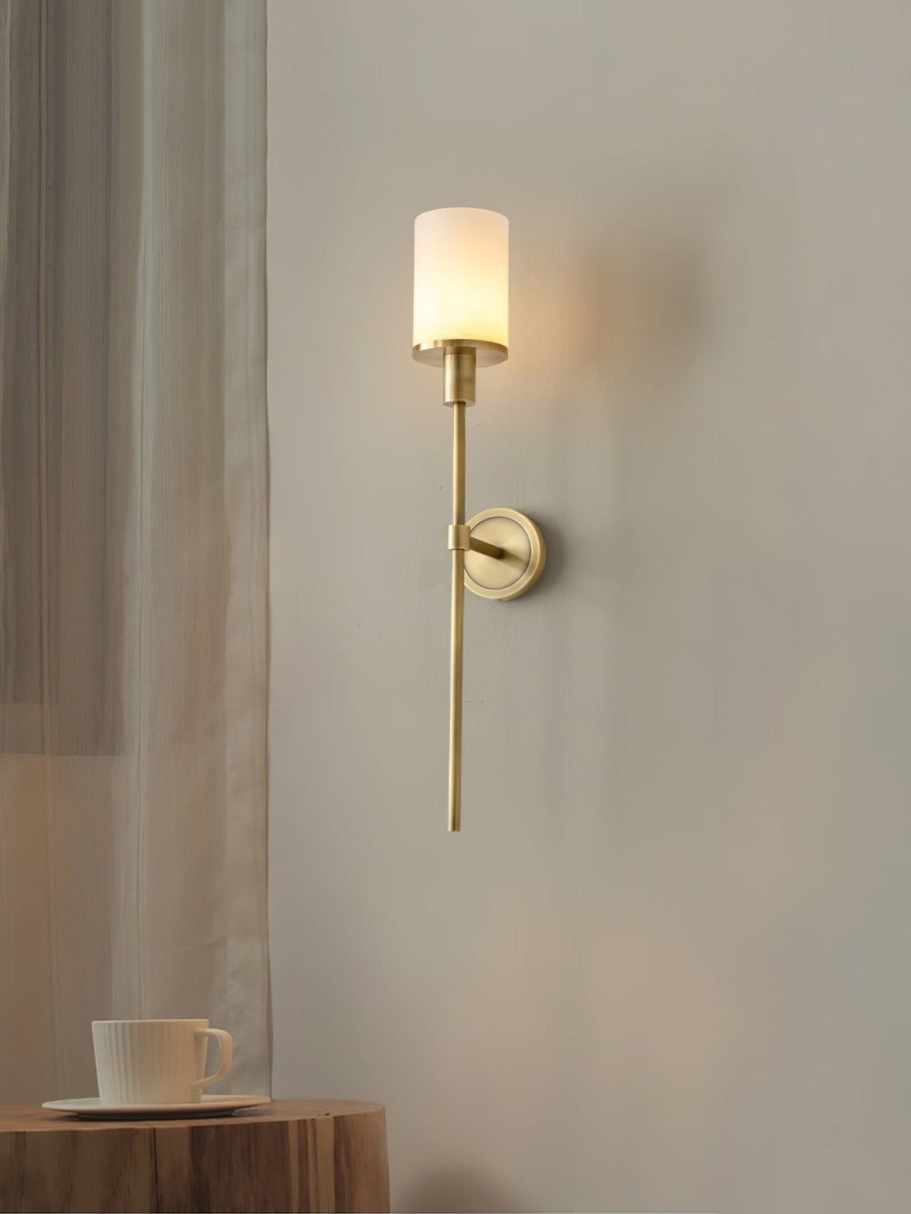 Clérachic brass alabaster sconce lit next to a sheer curtain, above a wooden stump table set with a cup