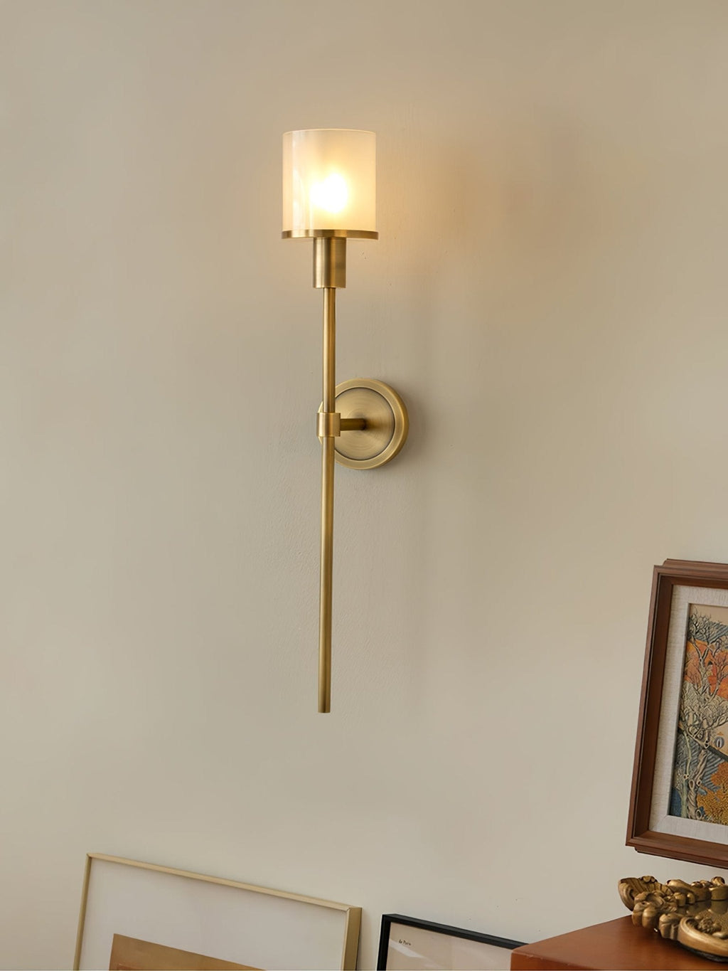 Clérachic brass alabaster sconce lit on a cream wall, with leaning picture frames and a wood dresser below
