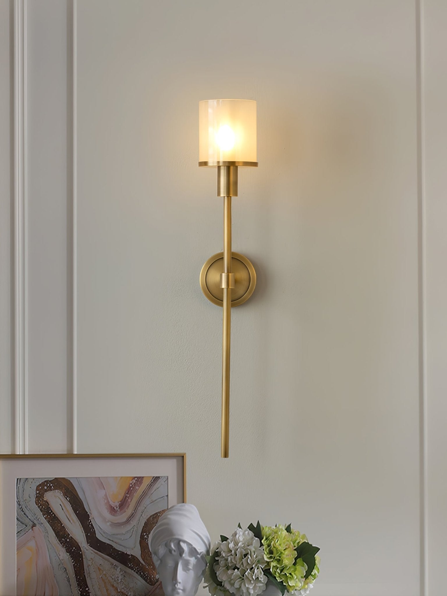Clérachic brass alabaster sconce lit on a panelled wall above framed art, a plaster bust and hydrangeas