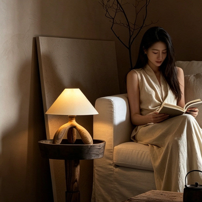 Beside an armchair where someone reads, this dark wood Clérachic lamp glows on a round side table in a dim room.